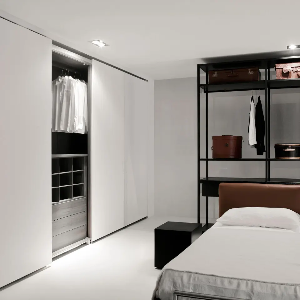 Camera da letto dai tono candidi, con armadio bianco sulla sinistra e armadio a giorno sulla parete di fondo, dietro al letto