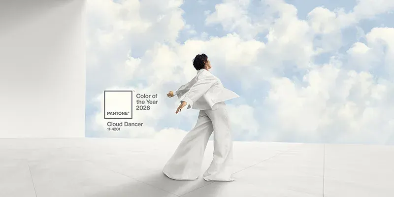 Visual del Pantone Color of the Year 2026 Cloud Dancer: a sinistra campione di colore bianco caldo con logo Pantone, a destra donna vestita di bianco che si muove davanti a un cielo azzurro con nuvole e box con scritta ‘Color of the Year 2026 – Cloud Dancer 11-4201