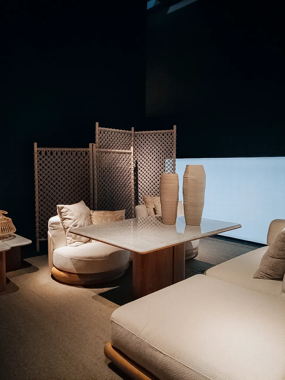 tavolo in marmo con vasi scultorei color sabbia, poltrona imbottita e paravento in vimini in un elegante ambiente fieristico